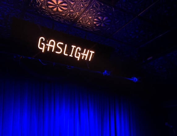 A dimly lit stage with the word 'GASLIGHT' in glowing white neon letters above. The backdrop features dark blue curtains, and the ceiling displays an ornate pattern. The overall atmosphere suggests a vintage or intimate performance space.