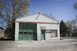A small, white-painted garage with a large green garage door and a smaller door, both made of weathered wood. There are signs that read 'AMOS Melba SERVICE' and 'INTERSTATE BATTERIES'. The building is surrounded by trees, with a clear blue sky.
