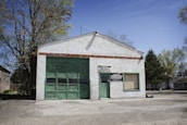 A small, white-painted garage with a large green garage door and a smaller door, both made of weathered wood. There are signs that read 'AMOS Melba SERVICE' and 'INTERSTATE BATTERIES'. The building is surrounded by trees, with a clear blue sky.