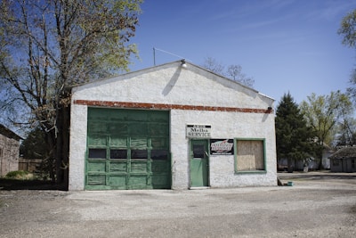 A small, white-painted garage with a large green garage door and a smaller door, both made of weathered wood. There are signs that read 'AMOS Melba SERVICE' and 'INTERSTATE BATTERIES'. The building is surrounded by trees, with a clear blue sky.