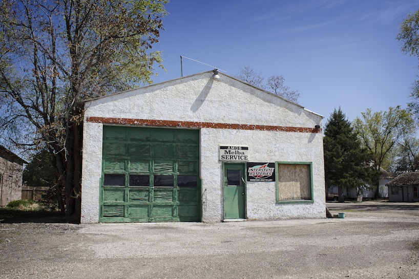 A small, white-painted garage with a large green garage door and a smaller door, both made of weathered wood. There are signs that read 'AMOS Melba SERVICE' and 'INTERSTATE BATTERIES'. The building is surrounded by trees, with a clear blue sky.