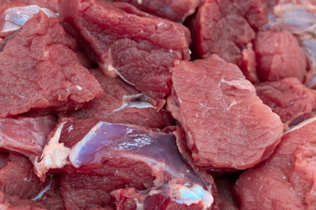 Raw pieces of red meat with visible marbling and texture are closely placed together, displaying pink and red hues with some surface moisture.