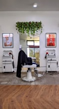 A modern barbershop setup includes a central barber chair with a black cloth draped over it. Two white workstations are placed symmetrically on either side, each equipped with tools and supplies. Above the mirror behind the chair, there is a lush arrangement of hanging green plants. Two framed posters flank the mirror, adding a touch of style to the setting. The hardwood floor and bright lighting contribute to a clean and inviting atmosphere.