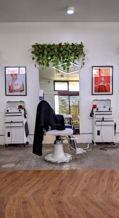 A modern barbershop setup includes a central barber chair with a black cloth draped over it. Two white workstations are placed symmetrically on either side, each equipped with tools and supplies. Above the mirror behind the chair, there is a lush arrangement of hanging green plants. Two framed posters flank the mirror, adding a touch of style to the setting. The hardwood floor and bright lighting contribute to a clean and inviting atmosphere.