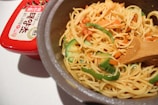 A pot filled with cooked noodles mixed with sliced green bell peppers and carrots, accompanied by a wooden spatula. Beside the pot, there is a red container with Korean writing on the label.