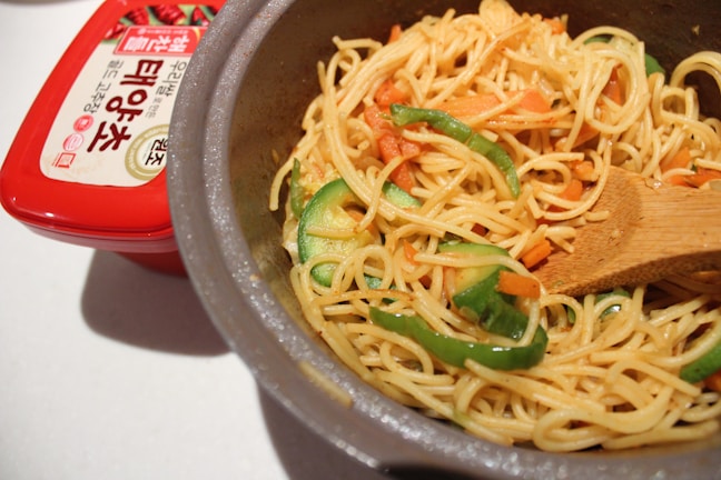 A pot filled with cooked noodles mixed with sliced green bell peppers and carrots, accompanied by a wooden spatula. Beside the pot, there is a red container with Korean writing on the label.