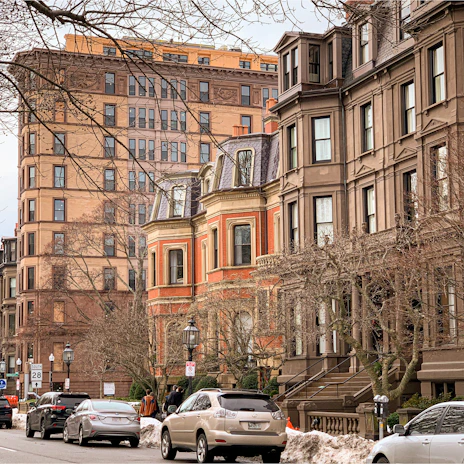 A street view featuring a line of brownstone buildings and an orange brick building with large windows. The architecture is detailed and historic. Several cars are parked on the street, and there are bare trees and street lamps. Snow is piled along the sidewalks, indicating a winter setting.