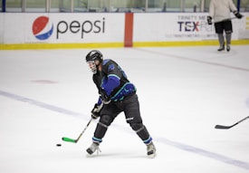 A hockey player in full gear, including a helmet and pads, skates across an ice rink while controlling a puck with their stick. The player is wearing a dark uniform with bright blue and green accents. Other players are visible in the background, and advertisements are displayed on the rink's boards.