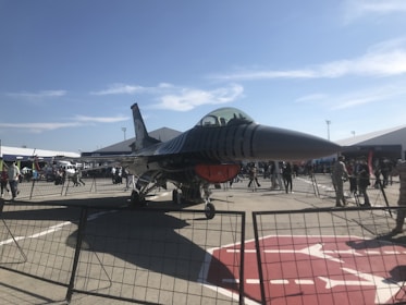 A modern military jet is displayed on an outdoor tarmac surrounded by a fence. Several people can be seen observing the aircraft, which is positioned under a clear blue sky with scattered clouds. Tents and additional vehicles are visible in the background, suggesting an airshow or exhibition setting.