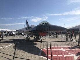 A modern military jet is displayed on an outdoor tarmac surrounded by a fence. Several people can be seen observing the aircraft, which is positioned under a clear blue sky with scattered clouds. Tents and additional vehicles are visible in the background, suggesting an airshow or exhibition setting.