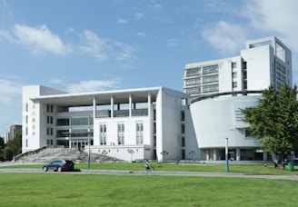 A modern, multi-storey building with a white facade and large glass windows, featuring both linear and curved architectural elements. The foreground has a well-maintained grassy area with a car parked on a path and a cyclist riding past. Surrounding the building are additional green spaces and some trees.
