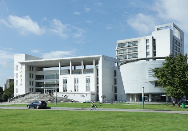 A modern, multi-storey building with a white facade and large glass windows, featuring both linear and curved architectural elements. The foreground has a well-maintained grassy area with a car parked on a path and a cyclist riding past. Surrounding the building are additional green spaces and some trees.