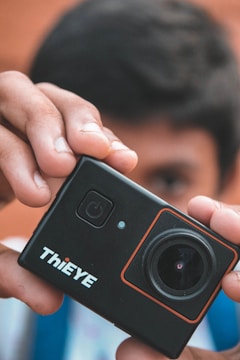 A close-up of a person's hands holding a compact, black action camera. The camera has a prominent lens and a power button visible on the front. The brand 'ThiEYE' is printed in white text on the camera's surface. A person's face is slightly blurred in the background, drawing attention to the camera in the foreground.