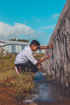 A person wearing a white shirt and dark pants is crouching near a small stream of water. They appear to be interacting with or examining a large concrete wall. The setting includes grassy vegetation and a blue sky with scattered clouds.