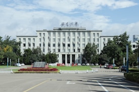 A large, multi-story institutional building with a symmetrical facade, surrounded by greenery and cars parked in front. The architecture is classic, with large windows and a central entrance. A well-maintained garden with red flowers is visible in the foreground.