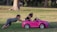 A young child is seated in a pink toy convertible car, holding something in their hands. Another child is playfully pushing the car from behind, using both hands. They are in a grassy park area with trees in the background, indicating a sunny outdoor setting.