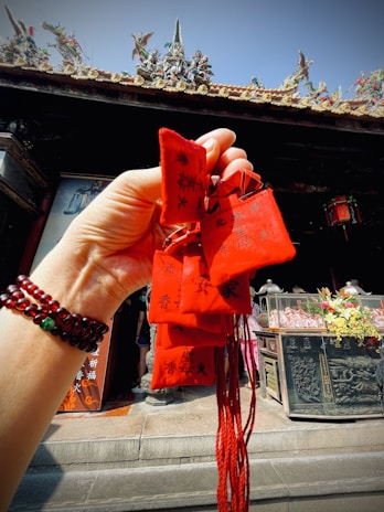 A hand wearing bead bracelets holds up several small red pouches with black Chinese characters. In the background, there is a traditional altar with offerings and decorative statues on a roof, under a clear blue sky.