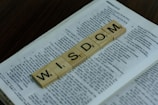 Wooden letter tiles spelling the word 'WISDOM' are placed on top of an open book. The book's pages contain text, indicating it might be a textbook or a piece of literature. The background is a dark, wooden surface.