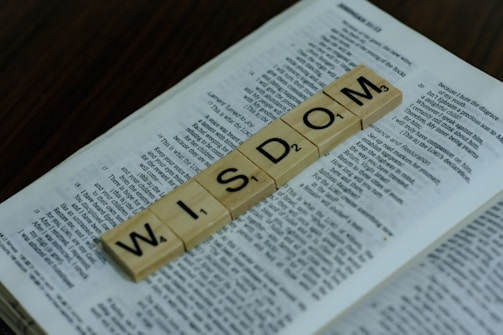 Wooden letter tiles spelling the word 'WISDOM' are placed on top of an open book. The book's pages contain text, indicating it might be a textbook or a piece of literature. The background is a dark, wooden surface.