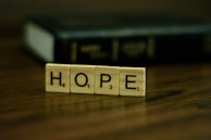 Wooden tiles with letters spelling out the word 'HOPE' are placed on a wooden surface. In the background, there is a blurred, dark-colored book.