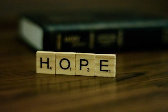 Wooden tiles with letters spelling out the word 'HOPE' are placed on a wooden surface. In the background, there is a blurred, dark-colored book.