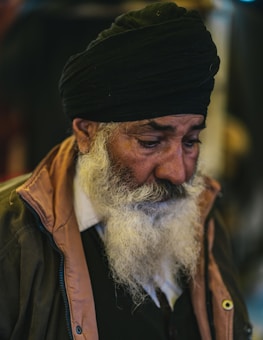 An elderly person with a long white beard wears a black turban and a brown jacket. Their expression appears contemplative, and they are looking downward.