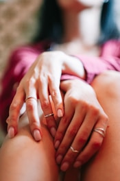 A close-up view focusing on hands resting on knees, with fingers adorned by multiple thin rings. The hands appear relaxed, creating a sense of calm and introspection. The background is softly blurred, directing attention toward the delicate details of the hands and jewelry.