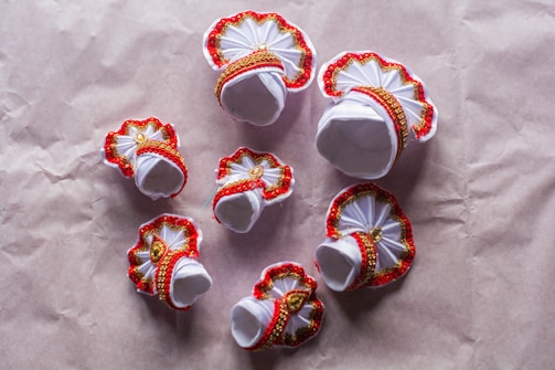 Several small, ornate decorations featuring white fabric adorned with red trim and gold embellishments. These appear to be handcrafted items set against a plain brownish background, possibly paper or fabric.