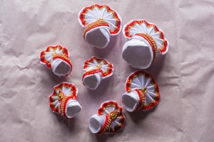 Several small, ornate decorations featuring white fabric adorned with red trim and gold embellishments. These appear to be handcrafted items set against a plain brownish background, possibly paper or fabric.