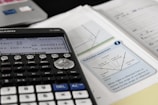 A scientific calculator is placed on top of a desk alongside a textbook and some notes. The calculator display shows some trigonometric calculations. The textbook page features a diagram explaining the concept of reflection, with terms like angle of incidence and angle of reflection. The surroundings contain other academic materials, suggesting a study environment.
