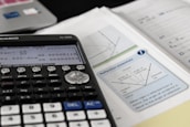 A scientific calculator is placed on top of a desk alongside a textbook and some notes. The calculator display shows some trigonometric calculations. The textbook page features a diagram explaining the concept of reflection, with terms like angle of incidence and angle of reflection. The surroundings contain other academic materials, suggesting a study environment.