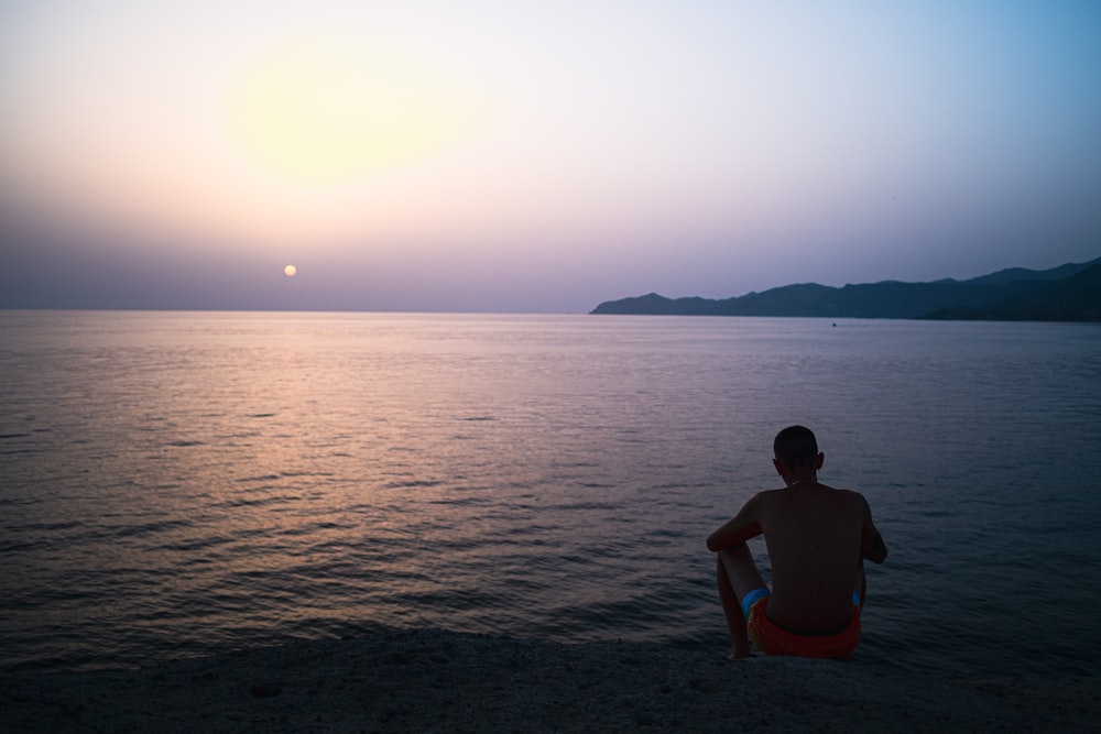 um homem sentado em uma praia observando o pôr do sol