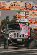 A rugged Jeep vehicle is parked prominently under an event banner that reads 'Afrodisia Get Ready for Adventure 2021.' The vehicle has a license plate reading 'LD 893' and displays a flag on the front. The background shows part of a modern building and trees.