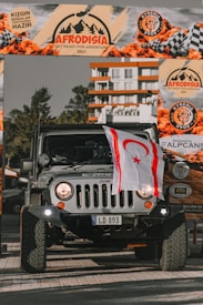 A rugged Jeep vehicle is parked prominently under an event banner that reads 'Afrodisia Get Ready for Adventure 2021.' The vehicle has a license plate reading 'LD 893' and displays a flag on the front. The background shows part of a modern building and trees.