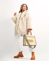 A person wearing a beige, plush overcoat paired with white trousers and tan ankle boots. They hold a stylish designer bag with a distinctive strap featuring a brand logo. The background is a soft off-white, which highlights the fashionable attire.