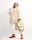 A person wearing a beige, plush overcoat paired with white trousers and tan ankle boots. They hold a stylish designer bag with a distinctive strap featuring a brand logo. The background is a soft off-white, which highlights the fashionable attire.