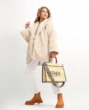 A person wearing a beige, plush overcoat paired with white trousers and tan ankle boots. They hold a stylish designer bag with a distinctive strap featuring a brand logo. The background is a soft off-white, which highlights the fashionable attire.
