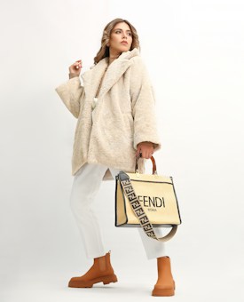 A person wearing a beige, plush overcoat paired with white trousers and tan ankle boots. They hold a stylish designer bag with a distinctive strap featuring a brand logo. The background is a soft off-white, which highlights the fashionable attire.