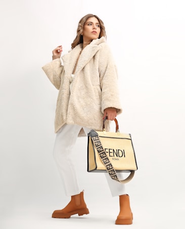 A person wearing a beige, plush overcoat paired with white trousers and tan ankle boots. They hold a stylish designer bag with a distinctive strap featuring a brand logo. The background is a soft off-white, which highlights the fashionable attire.