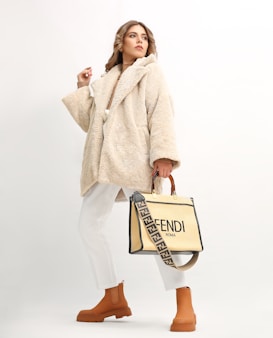 A person wearing a beige, plush overcoat paired with white trousers and tan ankle boots. They hold a stylish designer bag with a distinctive strap featuring a brand logo. The background is a soft off-white, which highlights the fashionable attire.
