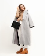 A stylish woman dressed in a long, gray coat over a beige turtleneck and white pants. She carries a quilted black handbag with a chain strap and wears brown ankle boots. Her long, wavy hair is down, and she gazes thoughtfully to the side against a light, neutral background.