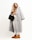 A stylish woman dressed in a long, gray coat over a beige turtleneck and white pants. She carries a quilted black handbag with a chain strap and wears brown ankle boots. Her long, wavy hair is down, and she gazes thoughtfully to the side against a light, neutral background.