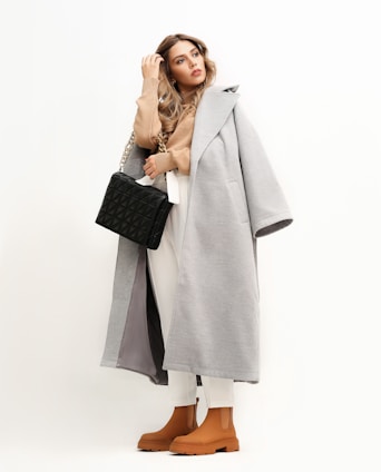 A stylish woman dressed in a long, gray coat over a beige turtleneck and white pants. She carries a quilted black handbag with a chain strap and wears brown ankle boots. Her long, wavy hair is down, and she gazes thoughtfully to the side against a light, neutral background.