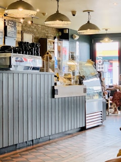 A cozy café interior featuring a coffee counter with an espresso machine and stacked paper cups. The dim lighting is provided by vintage-style pendant lights hanging from the ceiling. The décor includes a rustic brick wall, a wooden counter, and a display case filled with pastries or sandwiches. Customers are seen in the seating area near a window, creating a warm and inviting atmosphere.