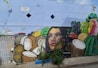 A vibrant mural on a wall features a woman's face surrounded by an assortment of fruits, including coconuts, bananas, grapes, oranges, and watermelon. The painting includes vivid colors and intricate details, with some greenery at the base.