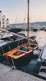 A small wooden sailboat is docked at a marina surrounded by other modern yachts. The water is calm, reflecting the boats and the light from the sky. Buildings are visible in the background, and the scene is set during twilight with soft lighting.