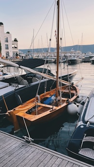 A small wooden sailboat is docked at a marina surrounded by other modern yachts. The water is calm, reflecting the boats and the light from the sky. Buildings are visible in the background, and the scene is set during twilight with soft lighting.