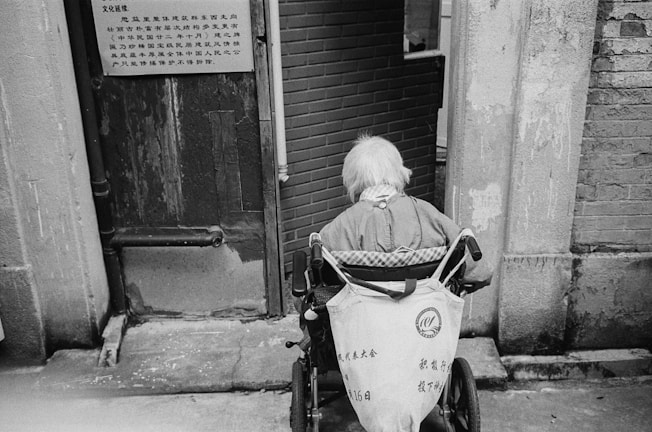 An elderly person with white hair sits in a wheelchair facing away from the camera. They are positioned near a worn doorway with textured walls and a brick interior visible beyond. A sign in an East Asian script is attached to the door nearby.
