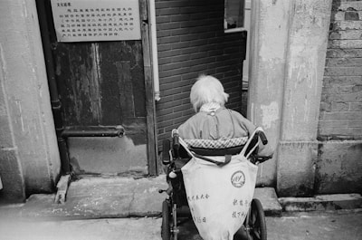 An elderly person with white hair sits in a wheelchair facing away from the camera. They are positioned near a worn doorway with textured walls and a brick interior visible beyond. A sign in an East Asian script is attached to the door nearby.