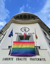 A building facade with the words 'HOTEL DE VILLE' and 'LIBERTE EGALITE FRATERNITE' displayed prominently. There are three flags on display: one is a rainbow flag, and the others include a European Union flag and a French national flag. The sky is blue with some clouds.
